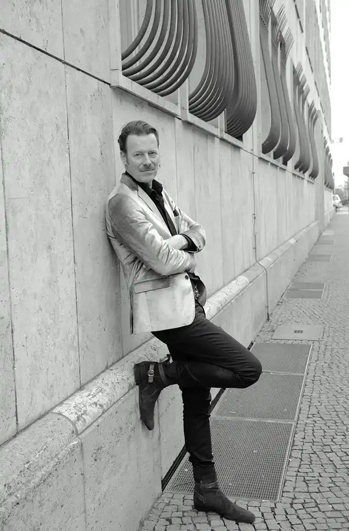 Christoph-Kamps Ein Mann in einem glänzenden Blazer lehnt an einer Steinmauer, ganz im Stil von Role Models Alter, die Arme selbstbewusst verschränkt.