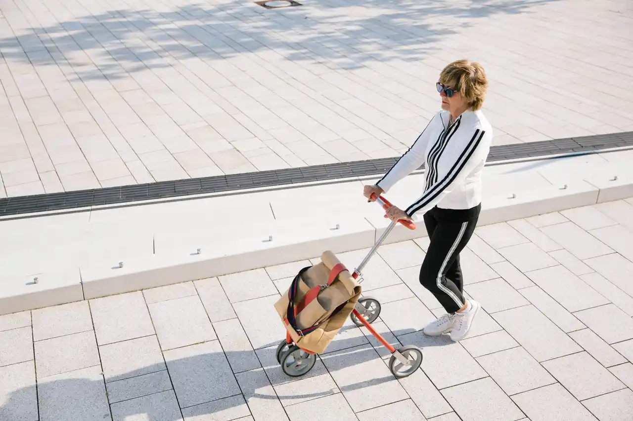 CityCaddy-in-HafenCity Person mit Sonnenbrille schiebt einen CityCaddy-Kinderwagen und umarmt selbstbestimmtes Altern auf einem sonnigen Spazierweg.