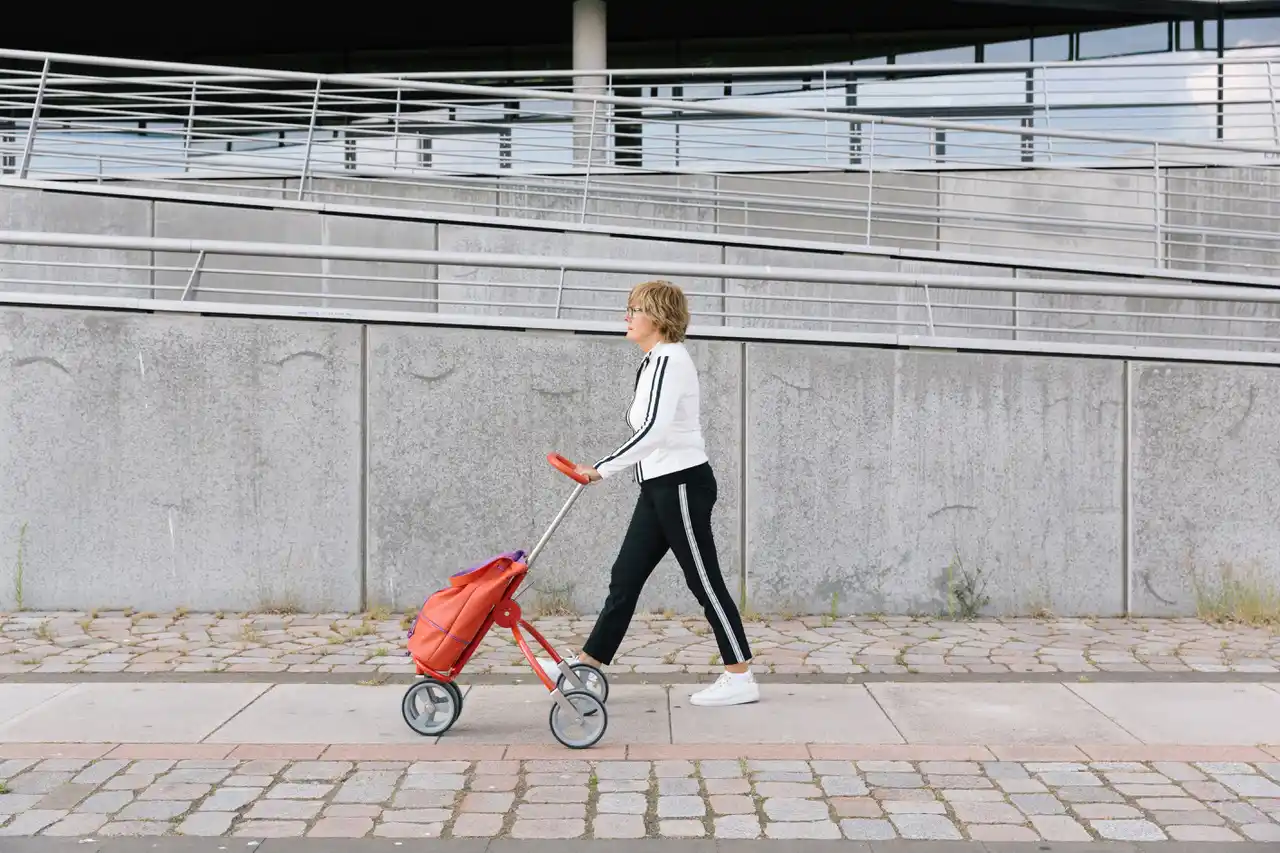 CityCaddy Eine Person in Sportkleidung geht auf einem Bürgersteig und schiebt einen roten CityCaddy-Wagen an einer Betonwand vorbei.