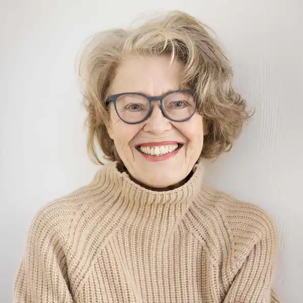 Ältere Frau mit grauen Haaren und Brille im beigen Pullover, die selbstbestimmtes Altern verkörpert, lächelt herzlich.