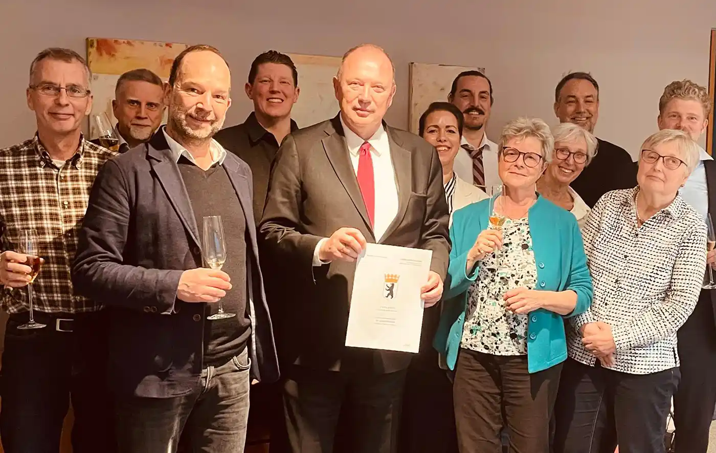 Notartermin-Baugruppe-Jardin-Hermsdorf Eine Gruppe posiert in einem Innenraum; einer im Anzug hält eine gerahmte Urkunde - die Baugemeinschaft Berlin ab 50 feiert gemeinsam.