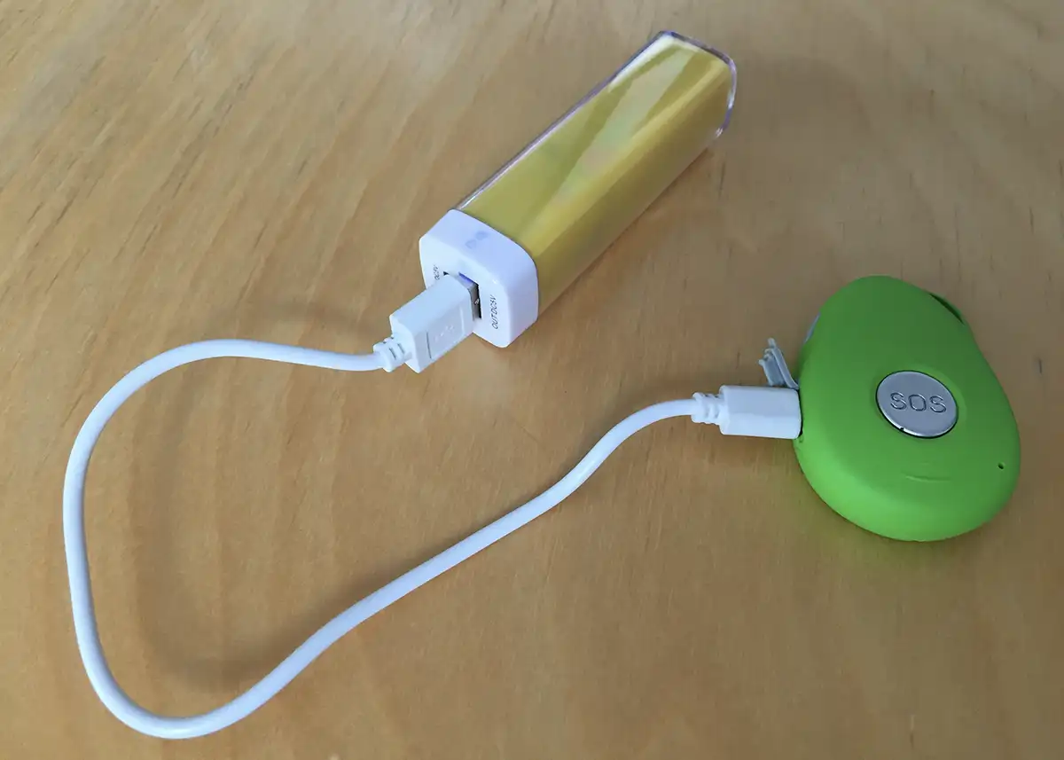 Eine gelbe Powerbank, die über ein weißes Kabel mit einem kleinen grünen SOS-Gerät auf einer Holzfläche verbunden ist.
