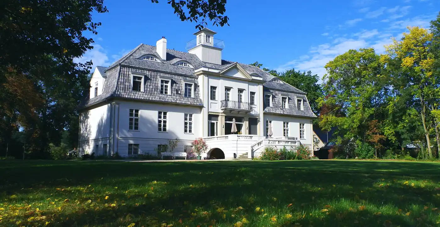 Schloss-Selchow_Christoph-Kamps Großes weißes Herrenhaus mit Säulen und Schornsteinen, ein Role Models Alter inmitten von Bäumen und einem gepflegten Rasen unter blauem Himmel.