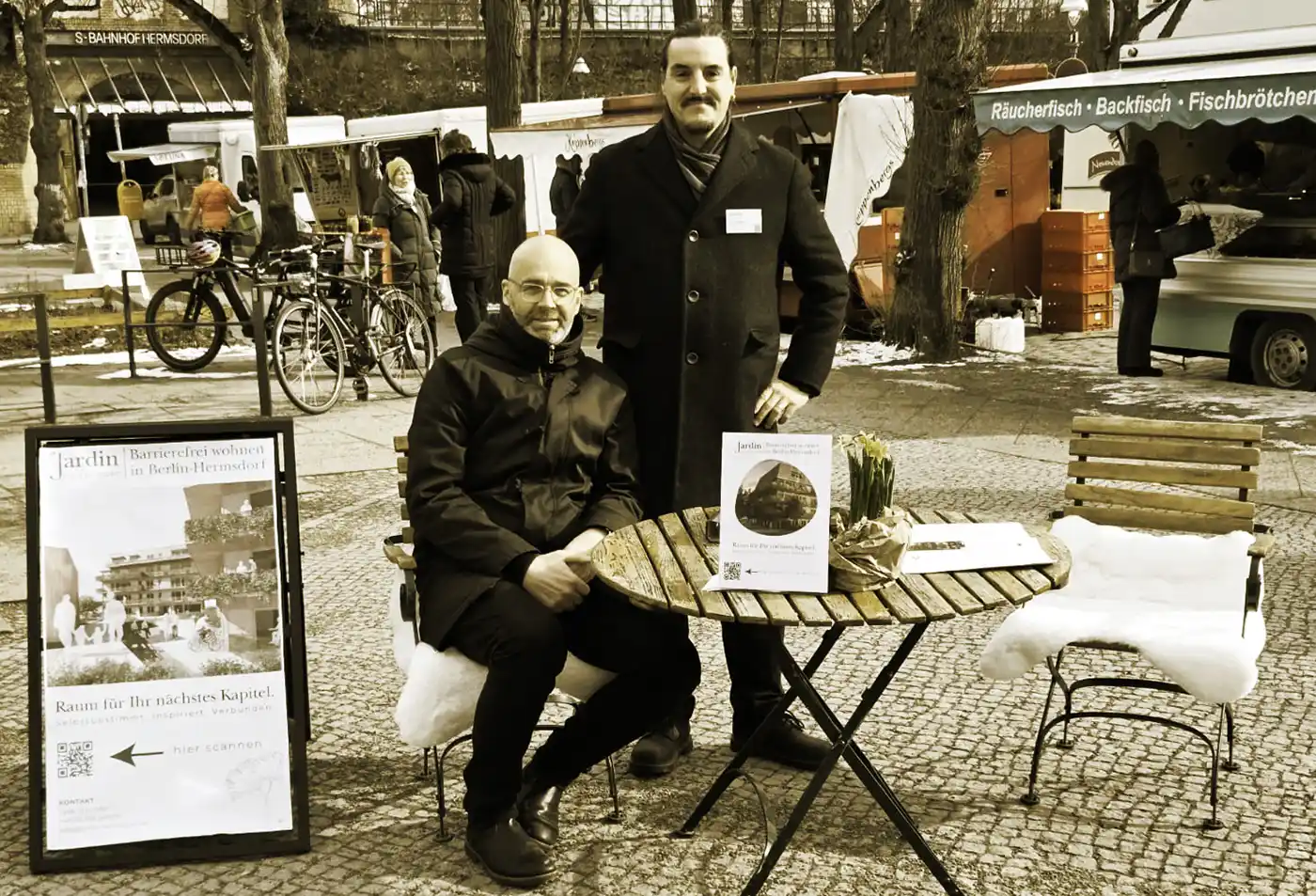Wochenmarkt-Jardin-Hermsdorf Zwei Männer an einem Tisch im Freien teilen sich die Baugemeinschaft Berlin ab 50 info auf einem belebten Markt mit Fahrrädern und Ständen.