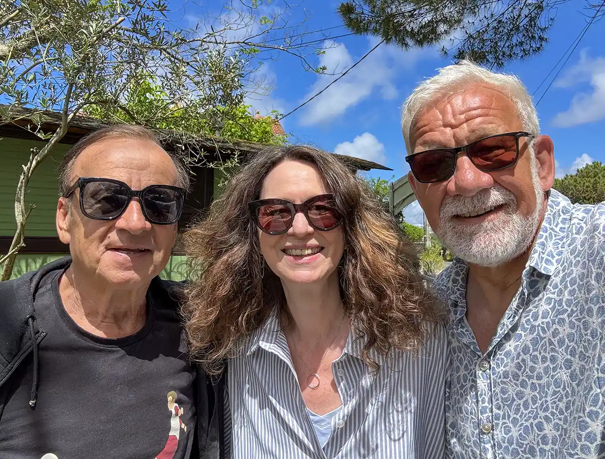 Gernot-Rohr-Claudia-Siegbert-Mattheis Drei Erwachsene mit Sonnenbrillen lächeln an einem sonnigen Tag im Freien mit Bäumen und einem Haus im Hintergrund.