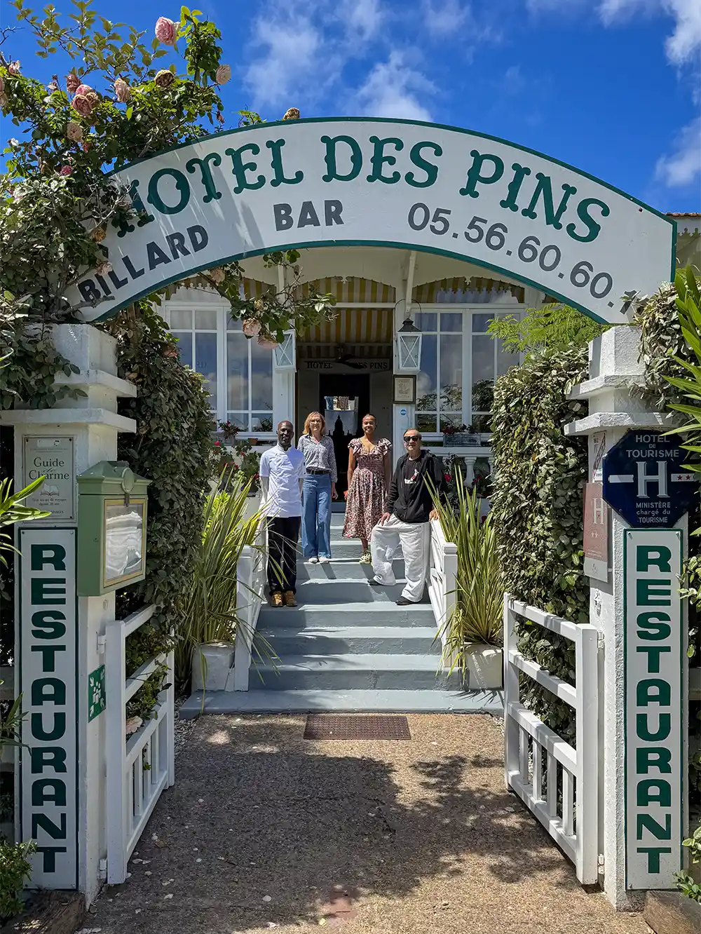 Gernot-Rohr-Hotel-des-Pins-Cap-Ferret Vier Personen stehen an einem sonnigen Tag auf den Stufen unter dem Eingangsschild des Hotel des Pins vor einem Restaurant.