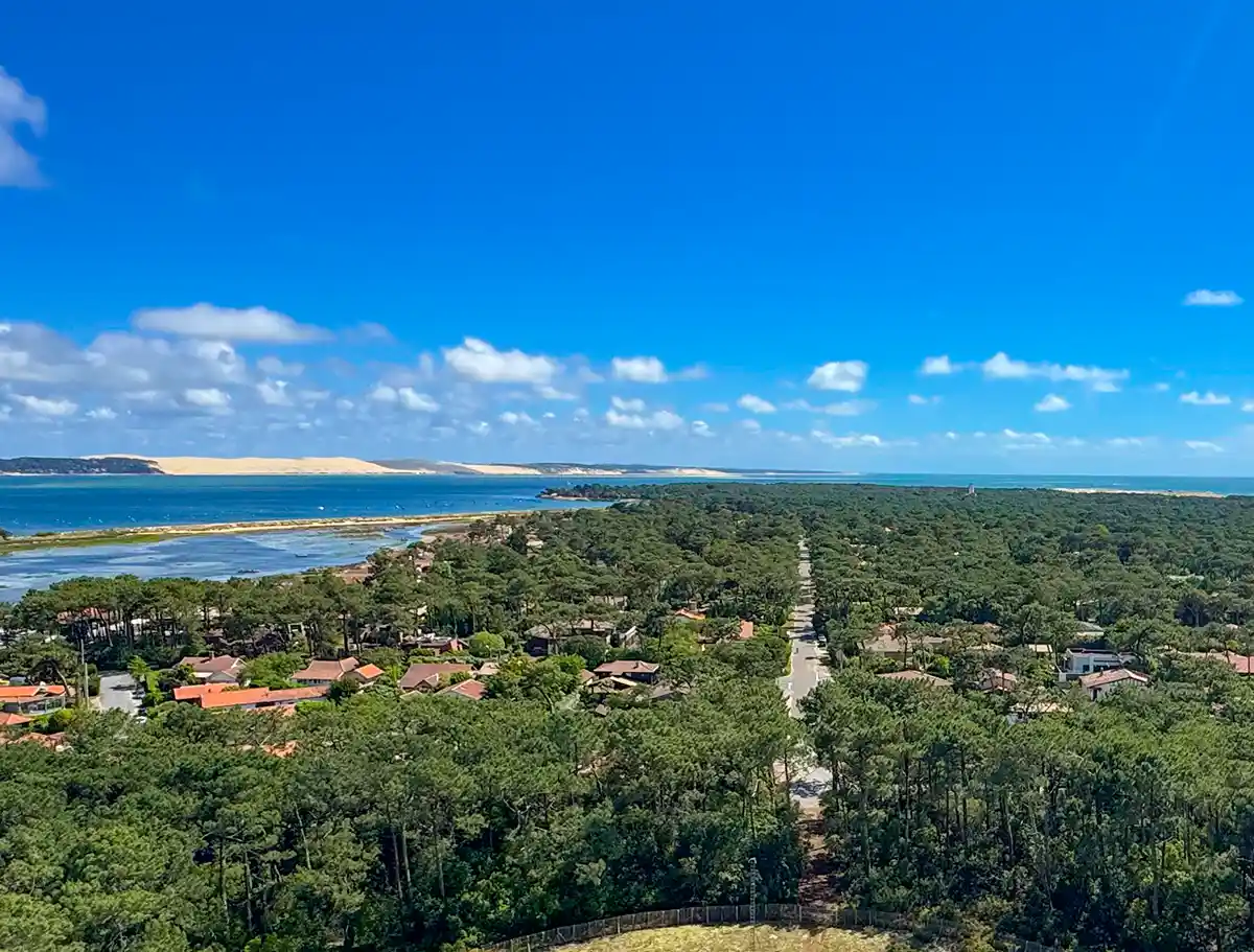 Cap-Ferret-Bassin-Arcachon Eine Küstenstadt mit von Bäumen gesäumten Häusern, einer Straße, die zum Meer führt, Sanddünen und einem strahlend blauen Himmel.
