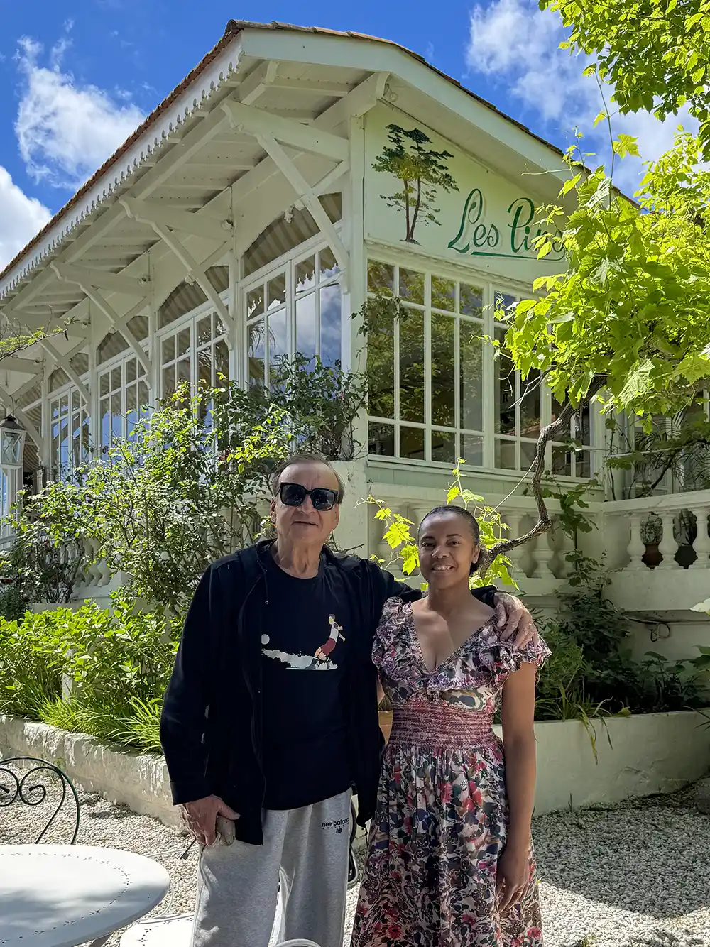 Cap-Ferret-Gernot-Rohr-Fabrina Ein Mann und eine Frau stehen an einem sonnigen Tag zusammen vor einer verglasten Veranda mit der Aufschrift Les Pins.