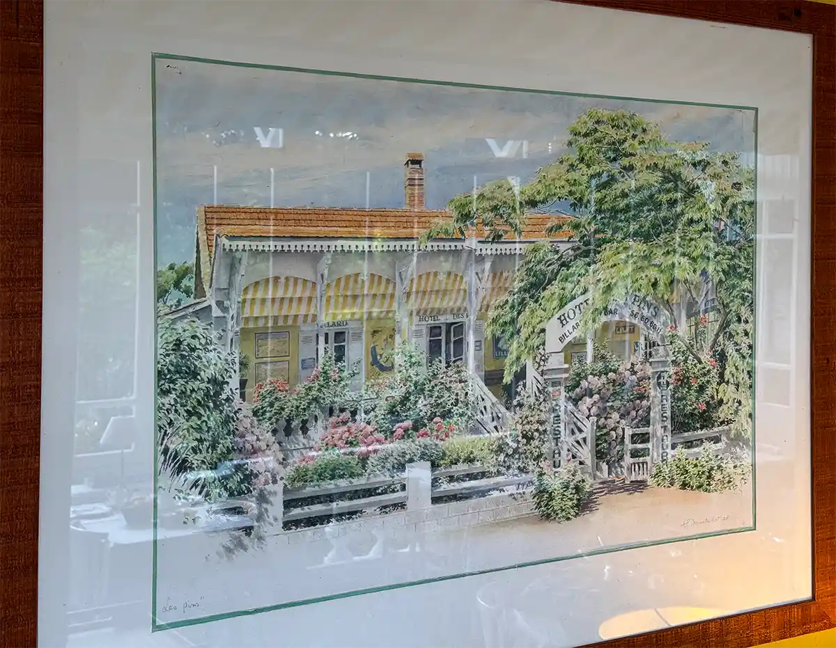 Cap-Ferret-Hotel-des-Pins-Aquarell-JP-Marladot-1998 Ein gerahmtes Gemälde, das ein gelbes Haus mit rotem Dach, einen Garten, Blumen und ein Schild mit der Aufschrift „Hotel“ am Eingang zeigt.
