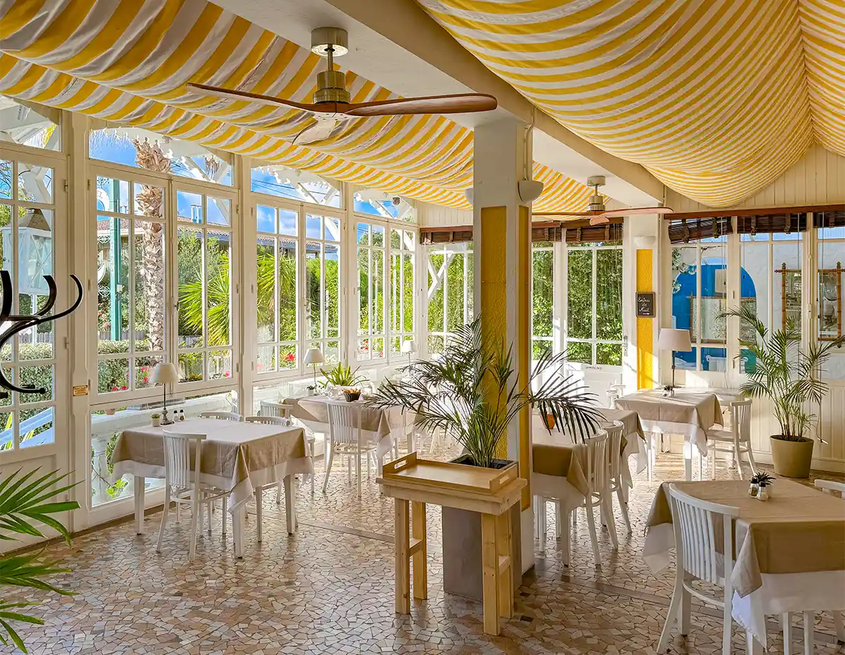 Cap-Ferret-Hotel-des-Pins-Restaurant Ein helles Restaurant mit großen Fenstern, einer gelb gestreiften Decke und gedeckten Tischen.