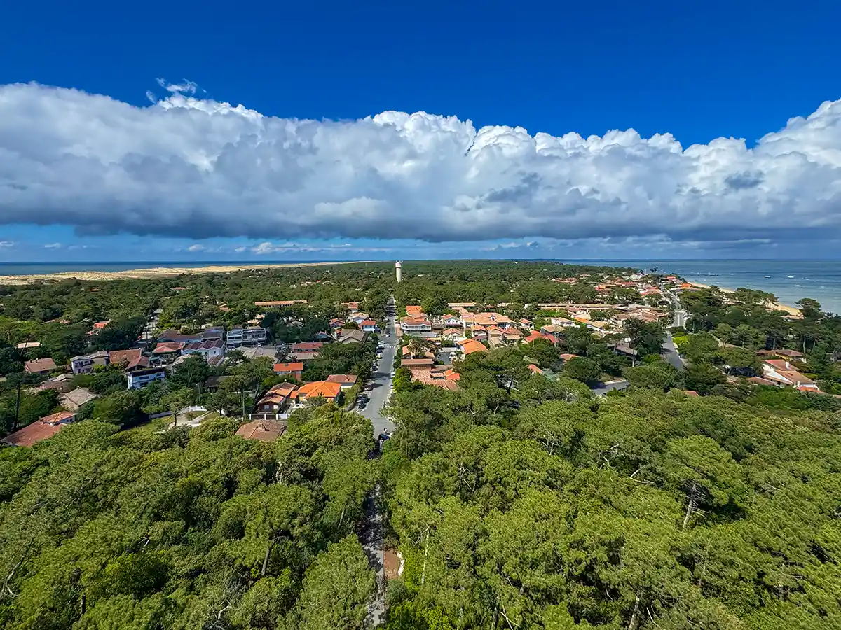 Cap-Ferret-Ozean-Arcachon Luftaufnahme einer Küstenstadt mit Häusern, dichtem Baumbestand, einem Leuchtturm und dem Meer unter einem blauen Himmel mit Wolken.