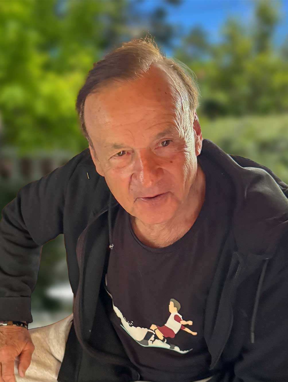 Gernot-Rohr-Hotel Älterer Mann in schwarzem Kapuzenpulli und grafischem T-Shirt, der im Freien sitzt, mit Grünzeug im Hintergrund.