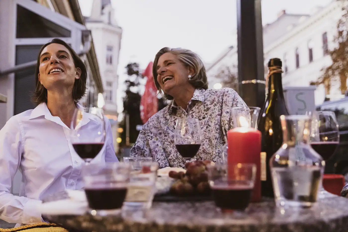 Yorokani_ Magnetkleidung_Frauen_Mood-02 Zwei Frauen in Magnetkleidung von Yorokani sitzen an einem gemütlichen Abend im Freien und lachen bei Wein, Kerzen und Snacks.