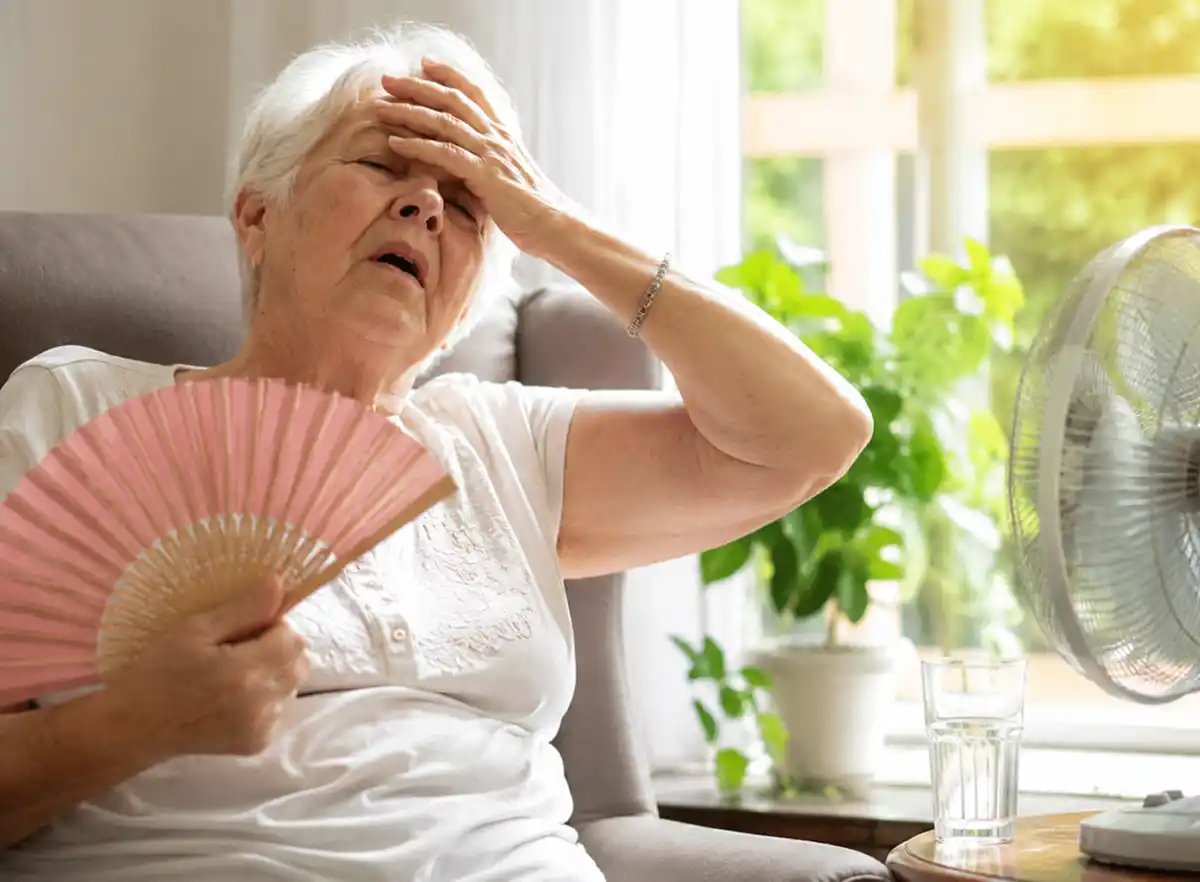 Hitze-aeltere-Menschen Eine ältere Frau, die sich Luft zufächelt und sich den Kopf hält, sitzt in der Nähe eines Ventilators und scheint in den Innenräumen unter Hitzestress zu leiden.