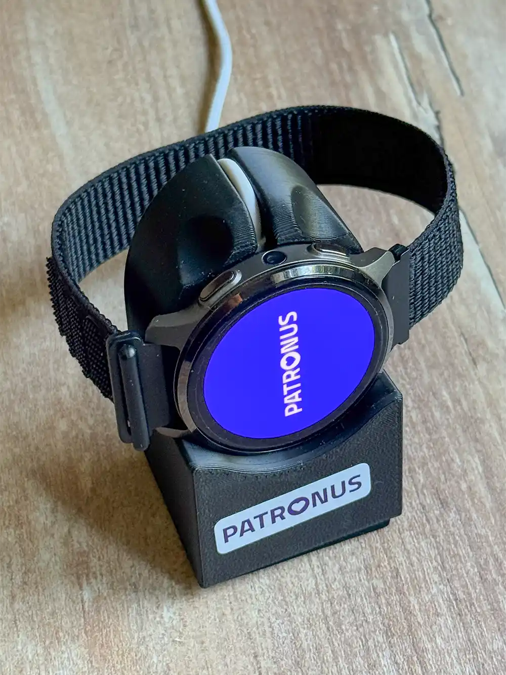 Patronus-Notfalluhr-Ladeschale Eine schwarze Smartwatch mit blauem Bildschirm zeigt „PATRONUS“ an, während sie auf einer beschrifteten Ladestation aufgeladen wird.