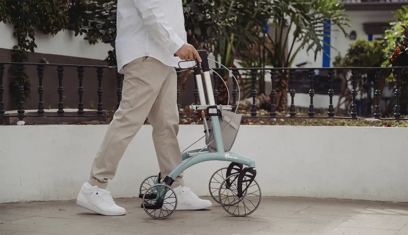Saljol_Rollator_Allrounder-Superlight-62 Eine Person, die die Freiheit auf vier Rädern genießt und mit einem blauen Rollator und leichter Kleidung im Freien spazieren geht.