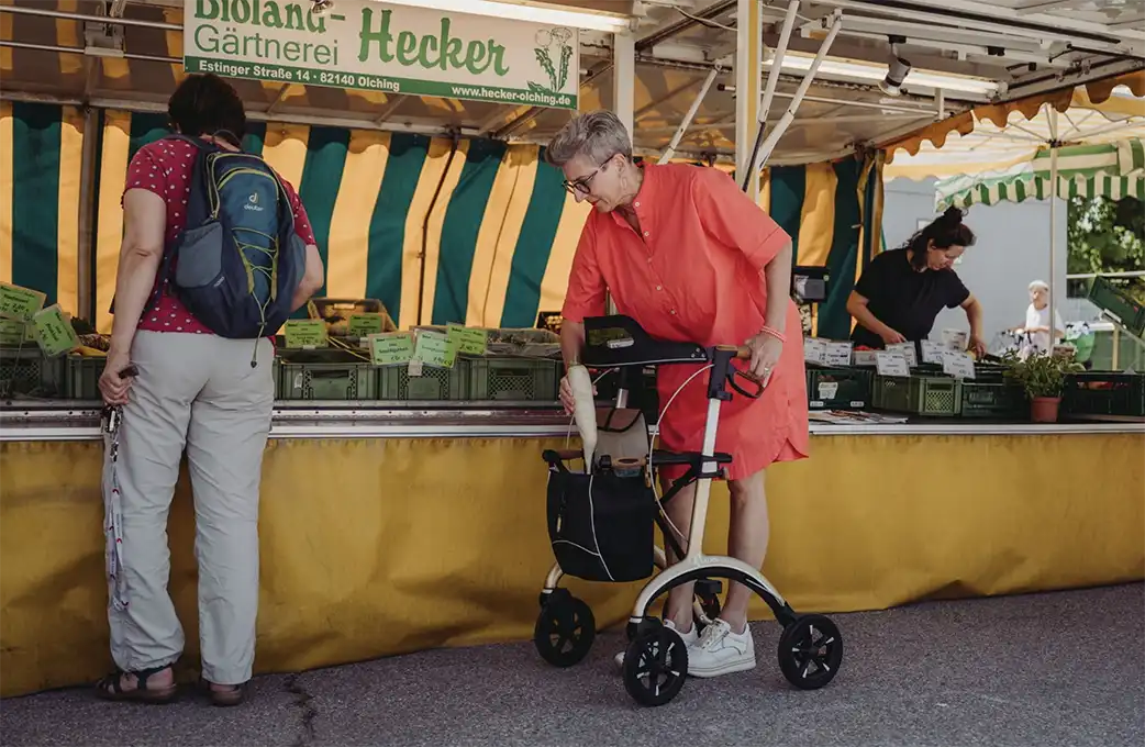Saljol_Rollator_Pure-Gold Eine ältere Frau in einem orangefarbenen Kleid benutzt einen Rollator, während sie auf einem Markt mit gelb gestreifter Markise einkauft.