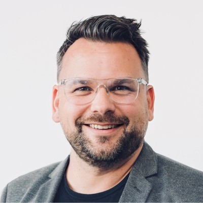 Ein lächelnder Mann in einem grauen Blazer und einer klaren Brille – das perfekte Bild für die Lifestyle-Kampagne „Wohnen im Ruhestand“.