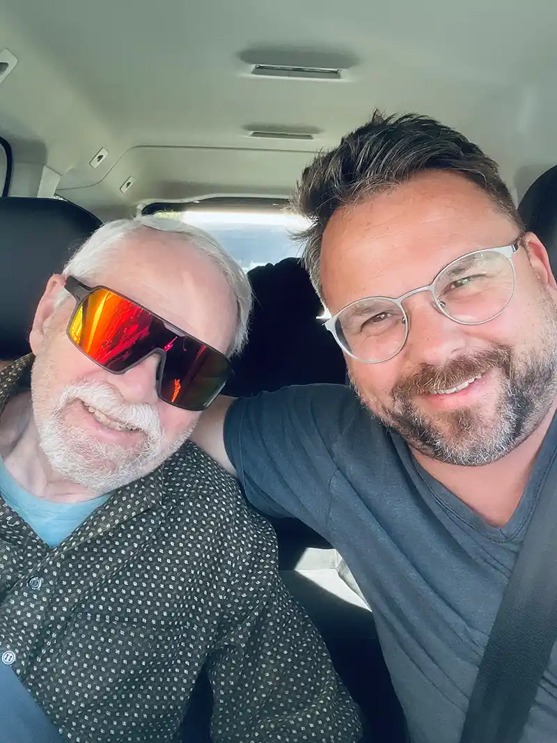 Tobias-Fruh_Roadtrip-Vater_Wohnen-Ruhestand Zwei Männer, die ihr Leben im Ruhestand genießen, sitzen lächelnd nebeneinander in einem Auto; der eine trägt eine Sonnenbrille, der andere eine Brille.