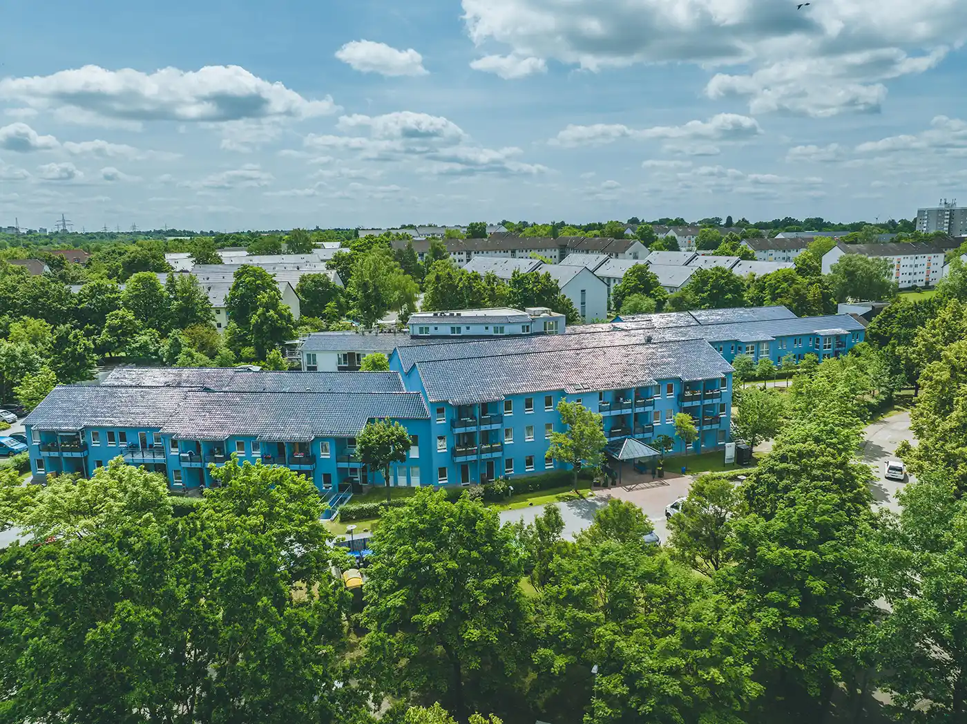 Tobias-Fruh_WIR-Wolfsburg-Fallersleben Luftaufnahme einer blauen Wohnanlage für Senioren, eingebettet zwischen Bäumen unter einem teilweise bewölkten Himmel.