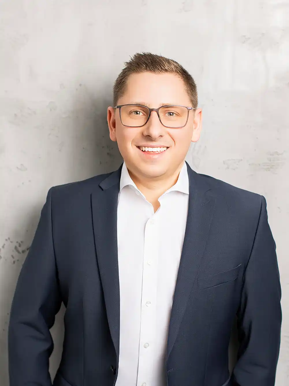 Oliver-Zirn Ein Mann mit Brille, der einen marineblauen Anzug und ein weißes Hemd trägt, steht vor einer hellgrauen, strukturierten Wand und lächelt.