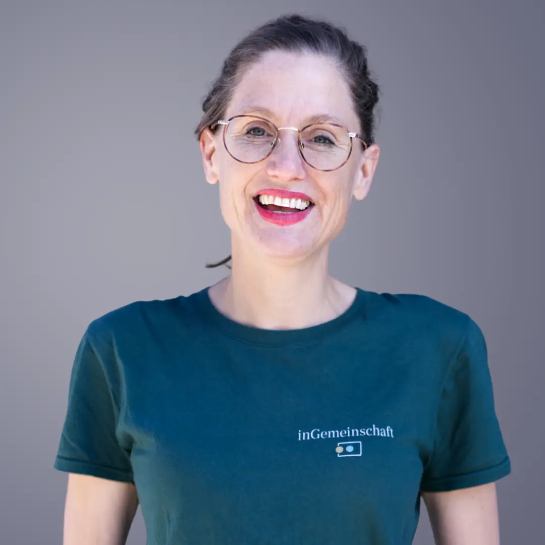 Eine Frau mit Brille, die in einem grünen „inGemeinschaft“-T-Shirt lächelt – perfekt für das Leben in einer Wohngemeinschaft ab 50.