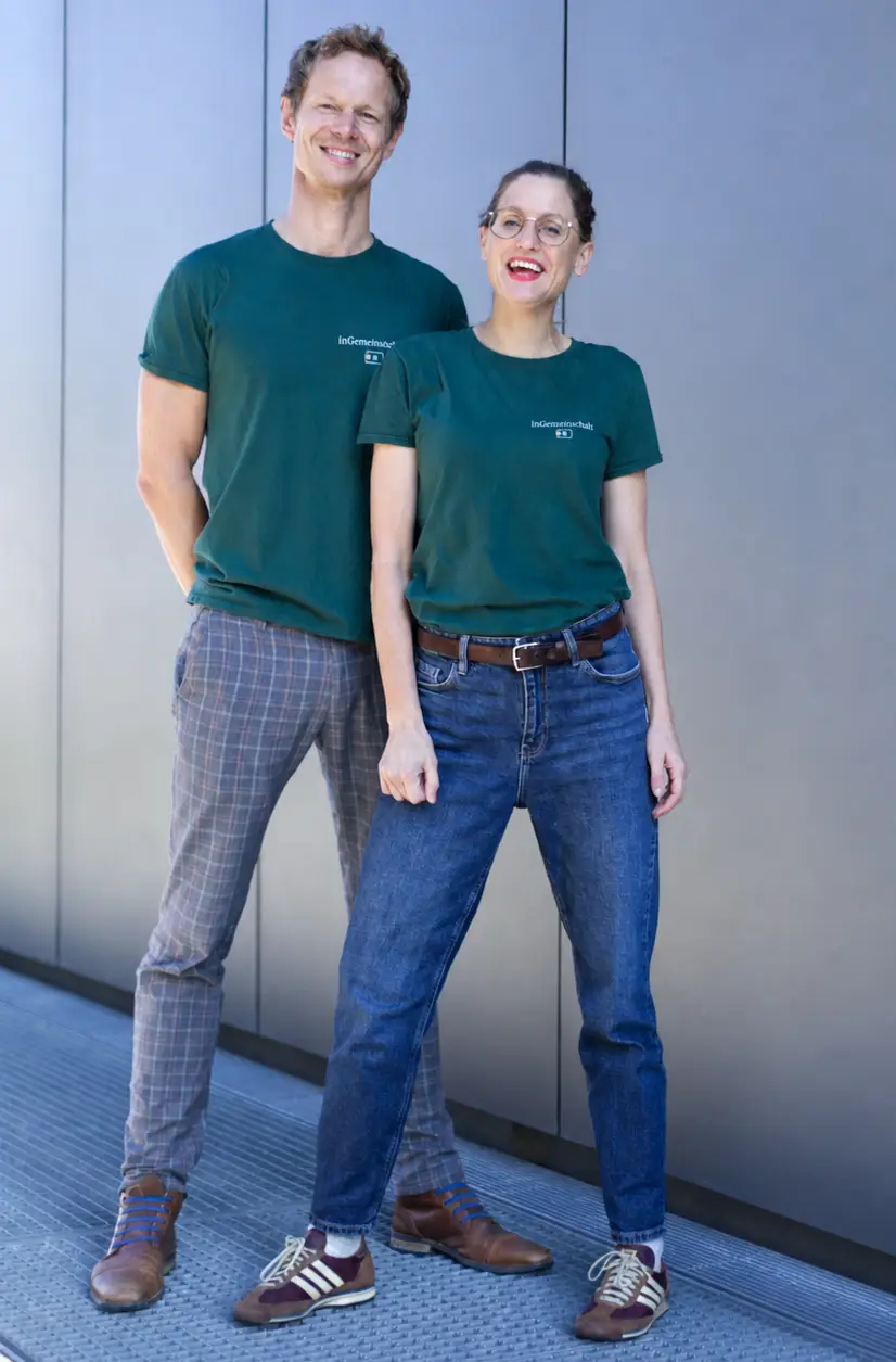 inGemeinschaft_Gruender_ Anne-Hufnagel_Philipp-Boegner Zwei Personen, die zu einer Wohngemeinschaft ab 50 gehören, stehen in grünen T-Shirts und mit Brillen vor einer grauen Wand nebeneinander.