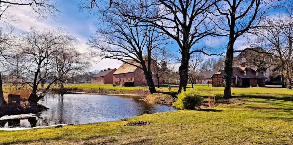 Ein Teich mit kahlen Bäumen und historischen Backsteingebäuden unter blauem Himmel in einer grasbewachsenen ländlichen Landschaft.