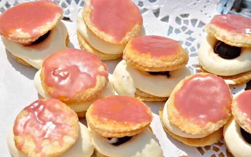 Gutleben-Buettnersdorf-Dessert Gebäck mit rosa Zuckerguss, Cremefüllung und dunklem Kern, angerichtet auf einem Tablett mit Spitzenmuster.