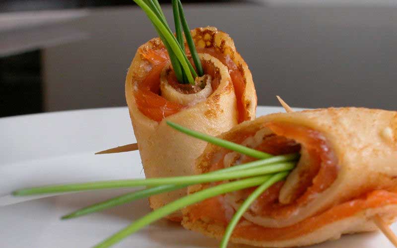 Gutleben-Buettnersdorf-Fingerfood Zwei gerollte Crêpes, gefüllt mit Räucherlachs und Schnittlauch, mit Zahnstochern fixiert, auf einem weißen Teller.